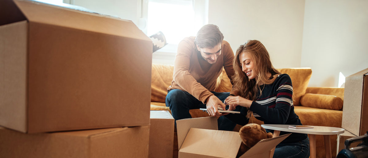 A man and woman sit on a couch surrounded by moving boxes, appearing relaxed despite the chaos of packing.