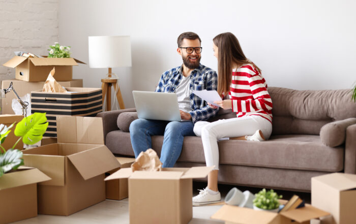 A couple sitting on a sofa surrounded by moving boxes, reviewing documents and using a laptop, illustrating how researching Removalist Insurance helps protect belongings during relocation.
