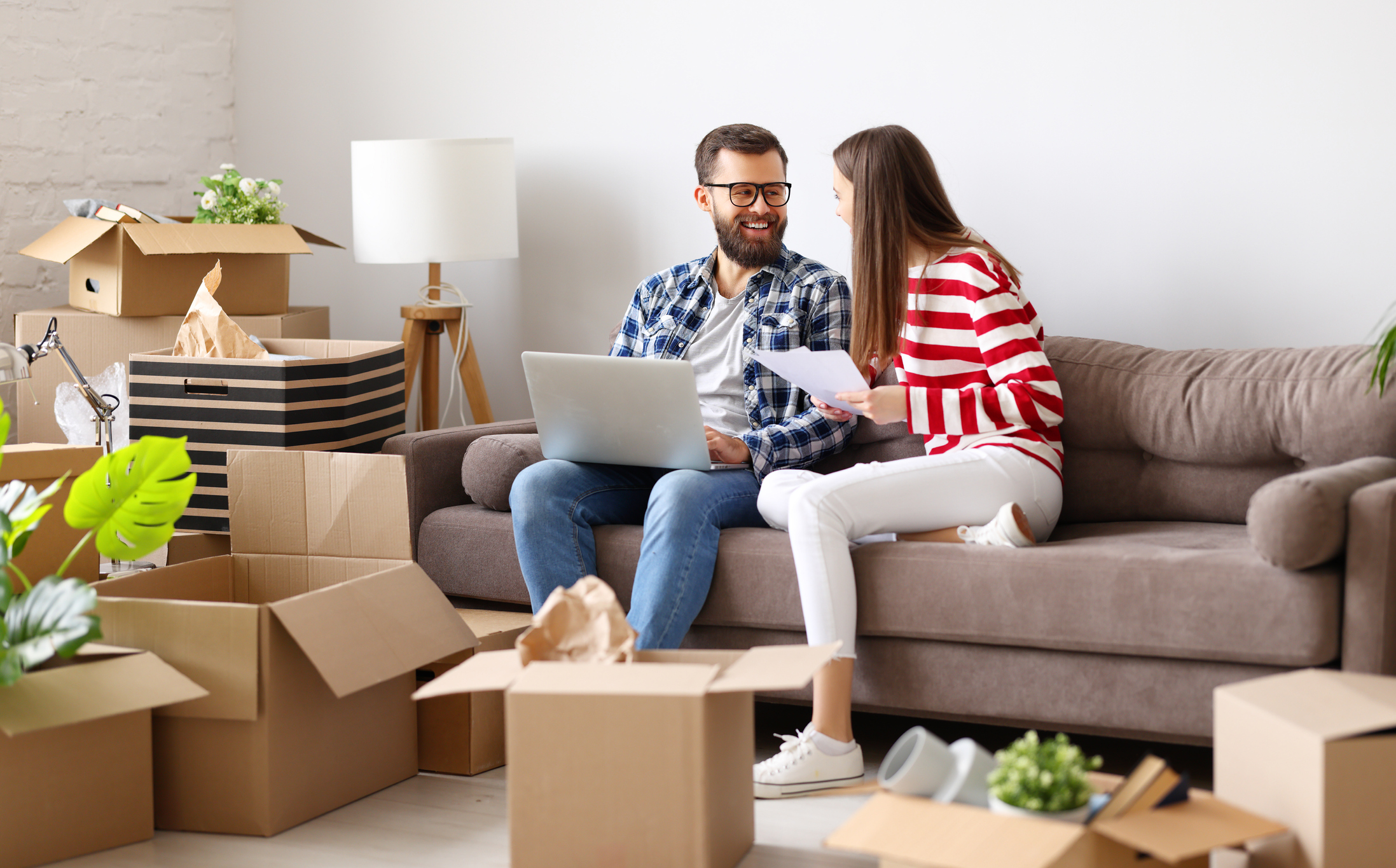 A couple sitting on a sofa surrounded by moving boxes, reviewing documents and using a laptop, illustrating how researching Removalist Insurance helps protect belongings during relocation.